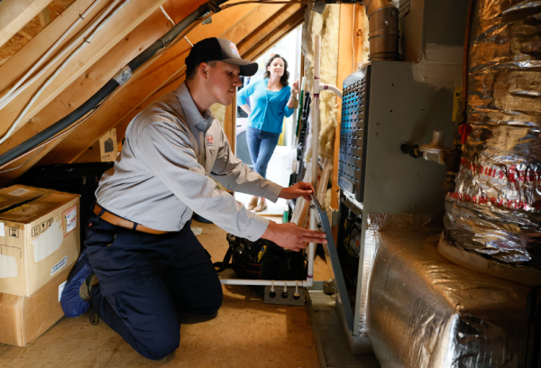 A Robert B. Payne Heating & Air technician kneeling in an attic to remove the service panel of a residential furnace while the homeowner looks on from the doorway