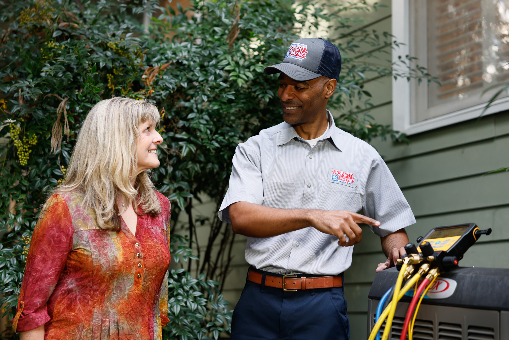 A Robert B. Payne Heating & Air technician pointing to a digital manifold gauge while explaining the performance of a Lennox outdoor AC unit to a female homeowner