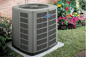 An outdoor American Standard air conditioning unit sits on a concrete pad in a yard next to a flower bed