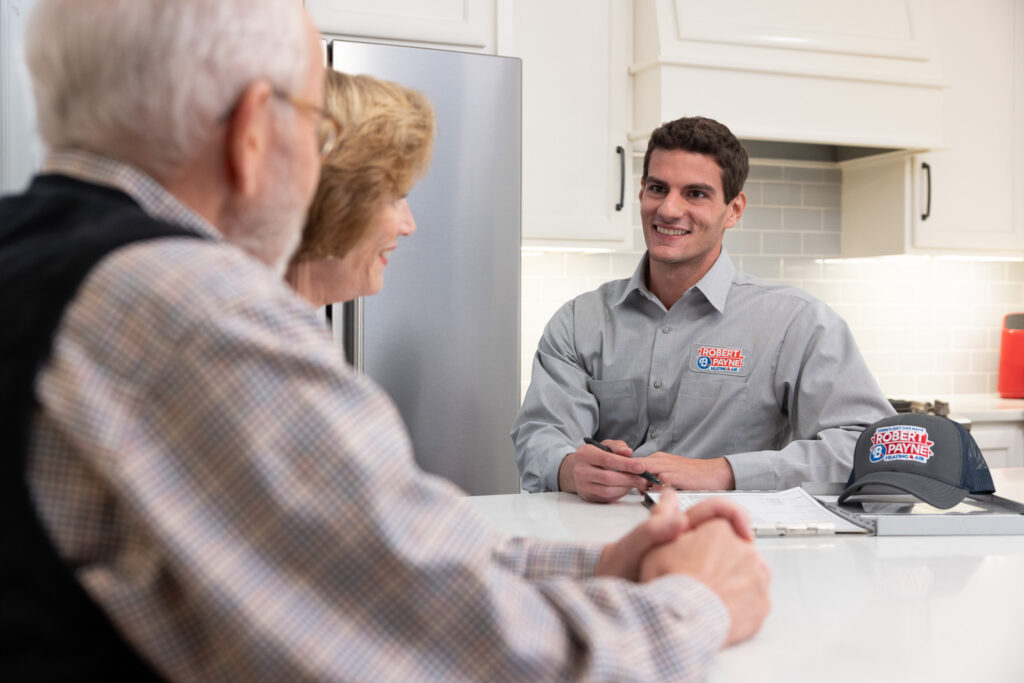 HVAC technician in a Robert B. Payne uniform speaks with homeowners | Robert B. Payne A smiling HVAC technician in a branded Robert B. Payne uniform sits at a kitchen counter across from an elderly couple and reviews paperwork with them