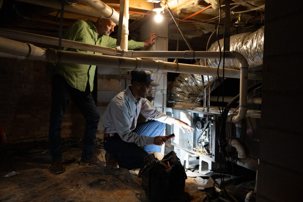 HVAC technician in a uniform uses a flashlight to inspect a furnace848 | Robert B. Payne An HVAC technician in a uniform uses a flashlight to inspect a furnace in a crawlspace while a man in a green shirt observes