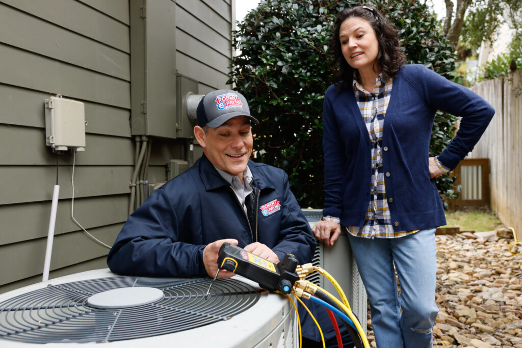 Robert B. Payne technician discusses an outdoor AC system with a homeowner | Robert B. Payne A Robert B. Payne technician uses a digital manifold gauge to service an outdoor air conditioning unit while speaking with a woman standing nearby