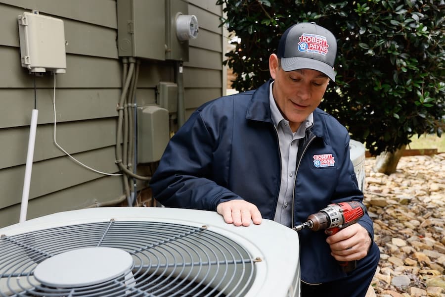 An HVAC technician in a branded jacket and cap uses a power drill to service an outdoor air conditioning condenser unit