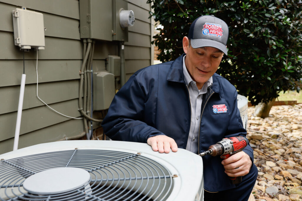 Robert B. Payne technician working on an AC unit | Robert B. Payne An HVAC technician in a branded jacket and cap uses a power drill to service an outdoor air conditioning condenser unit