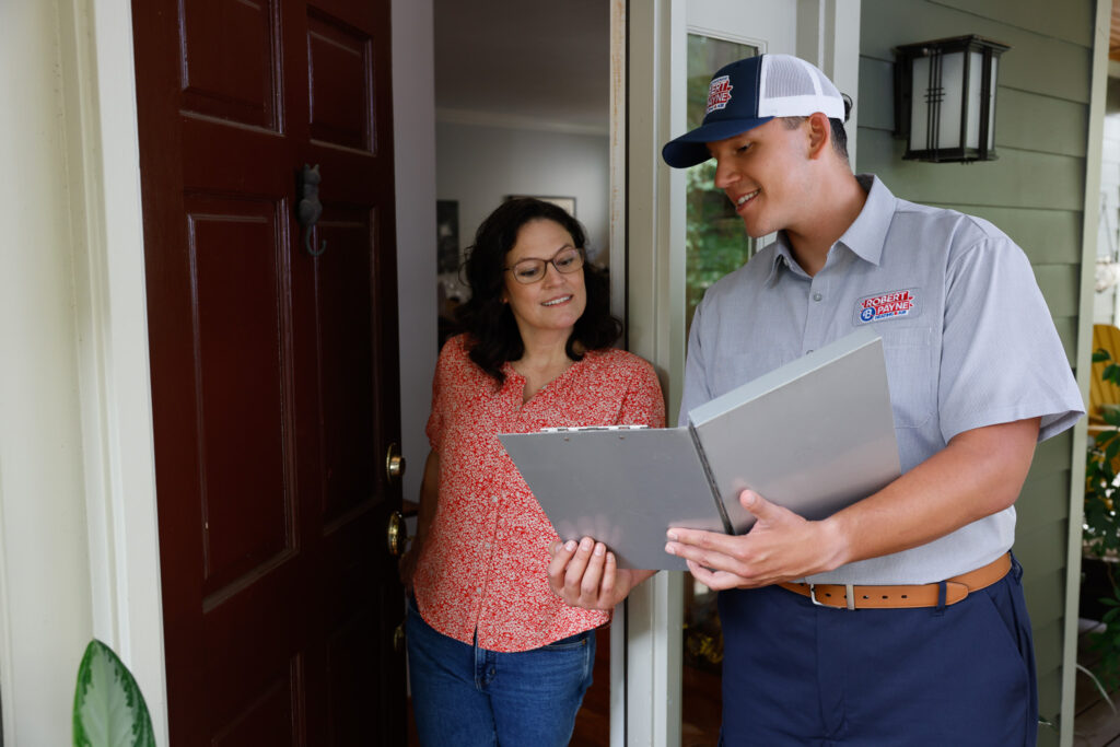 A smiling Robert B. Payne technician in a branded uniform reviews paperwork on a clipboard with a woman at the entrance of her home