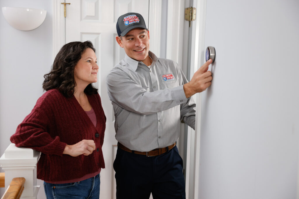 An HVAC technician in a Robert B. Payne branded uniform adjusts a round smart thermostat on a wall while a woman in a maroon cardigan observes
