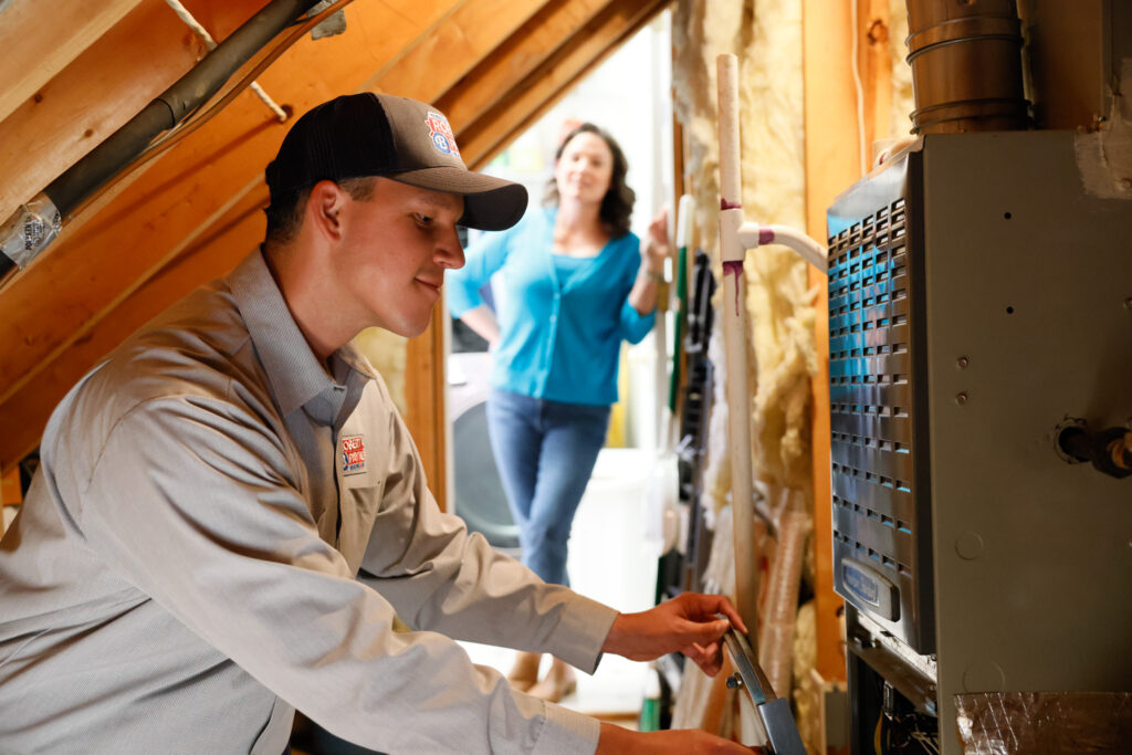 Robert B. Payne technician servicing an attic furnace | Robert B. Payne A Robert B. Payne technician in a branded uniform and cap performs maintenance on an attic furnace while a customer watches from the background