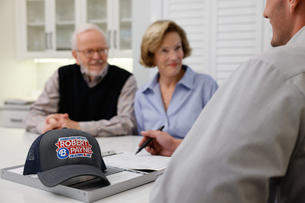 A Robert B. Payne technician reviews paperwork with an elderly couple at a kitchen counter, with a branded cap resting in the foreground