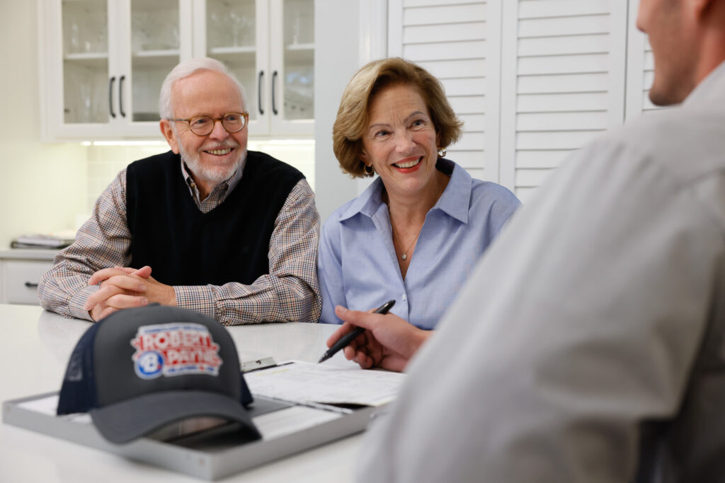 Robert B. Payne technician meeting with customers | Robert B. Payne A Robert B. Payne technician reviews paperwork with an elderly couple at a kitchen counter, with a branded cap resting in the foreground