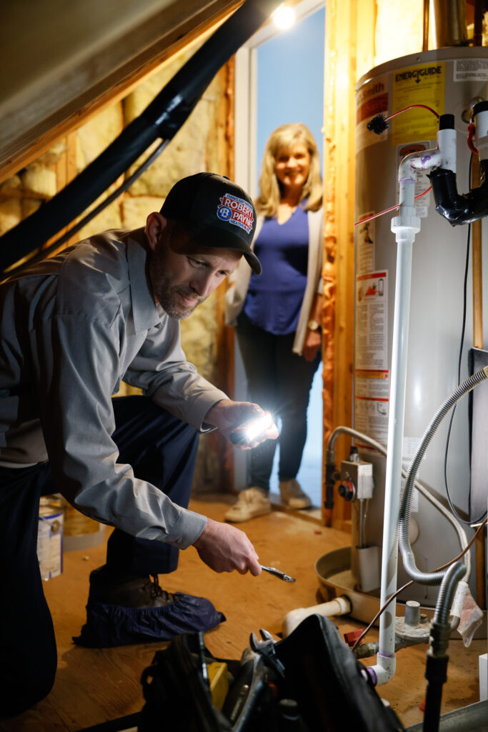 Robert B. Payne technician inspecting a water heater | Robert B. Payne A Robert B. Payne technician in a branded uniform uses a flashlight to inspect a water heater in an attic while a woman watches from the background