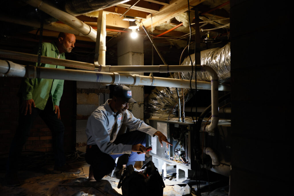 Robert B. Payne technician inspecting a furnace in a crawlspace | Robert B. Payne A Robert B. Payne technician uses a flashlight to inspect a furnace in a crawlspace while a man in a green shirt observes
