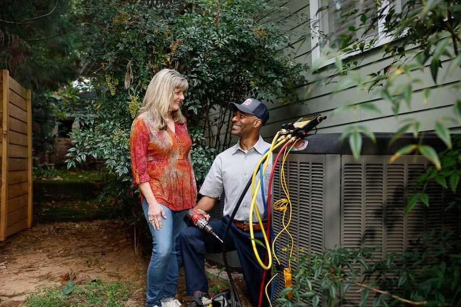 A smiling HVAC technician in a uniform and cap sits next to an outdoor air conditioning unit and speaks with a woman standing beside him