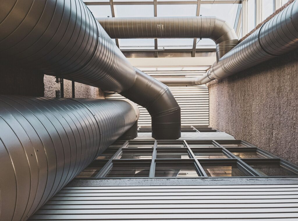 Large, cylindrical silver metal HVAC ducts run across a modern ceiling with skylights and glass panels